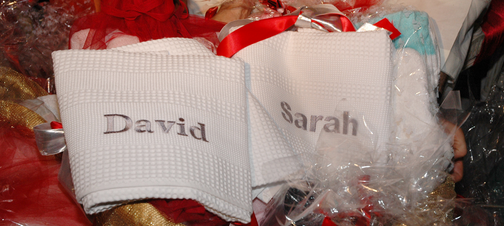 Serviettes personnalisées brodées avec les prénoms 'David' et 'Sarah', préparées pour un mariage, détails de décoration de table avec rubans rouges et dorés, ambiance festive et élégante.