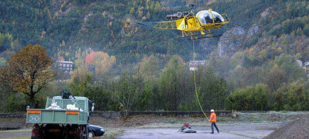 Hélicoptère jaune soulevant une charge par élingue en zone montagneuse, camion benne chargé de matériaux, chantier en extérieur, travaux de levage aérien, environnement forestier, opération logistique par hélicoptère.