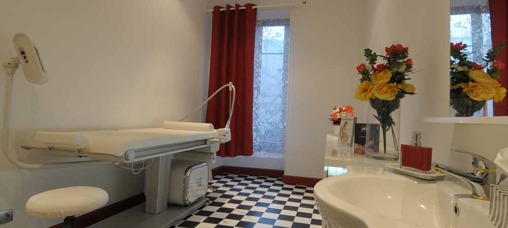 Salle de soin esthétique moderne avec table de traitement médicale, sol carrelé noir et blanc, lavabo élégant, bouquet de fleurs colorées, rideaux rouges, ambiance propre et lumineuse, institut de beauté ou cabinet médical.