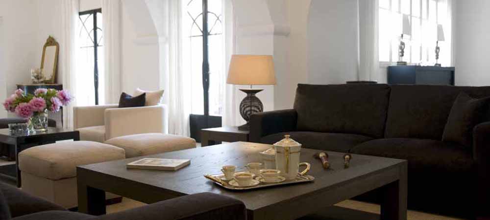 Salon élégant avec canapé gris, fauteuils beiges et table basse en bois, service à thé sur plateau, lampe design sur buffet, bouquet de fleurs fraîches, ambiance lumineuse grâce aux grandes fenêtres, décoration intérieure moderne et chaleureuse.