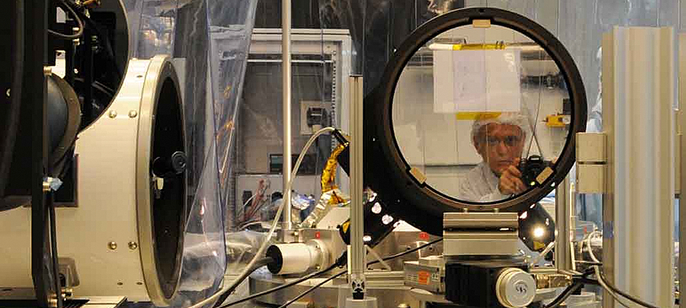 Photographe S-H en blouse de protection observant à travers un équipement optique de précision en laboratoire high-tech, recherche scientifique, instrumentation avancée, salle blanche, innovation technologique, matériel optique professionnel.