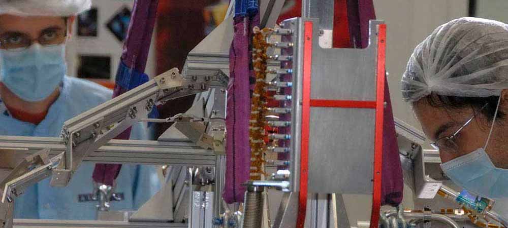 Deux techniciens en blouse et masque de protection travaillant en salle blanche sur un équipement électronique de haute précision, assemblage de composants technologiques, industrie high-tech, recherche et développement, environnement stérile, matériel scientifique, innovation technologique.